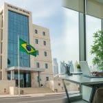 Funcionário administrativo trabalhando em escritório moderno na Embaixada do Brasil em Doha, com bandeira brasileira ao fundo, representando o processo seletivo para vaga de Auxiliar Administrativo em 2025.